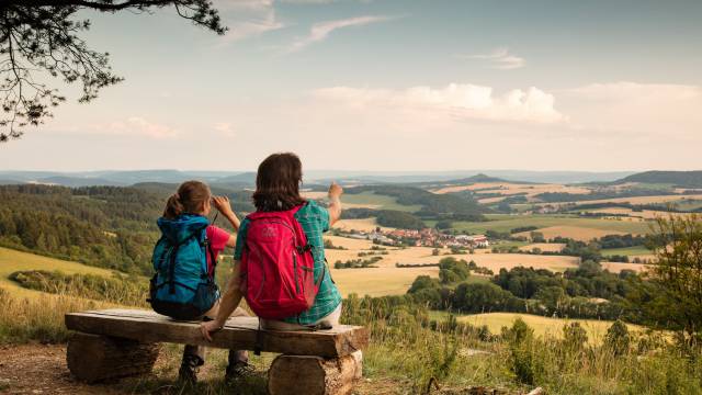 Zwei wandernde Personen mit Rucksäcken sitzen auf einer Bank und blicken über die weitläufige Hügellandschaft im Eichsfeld.