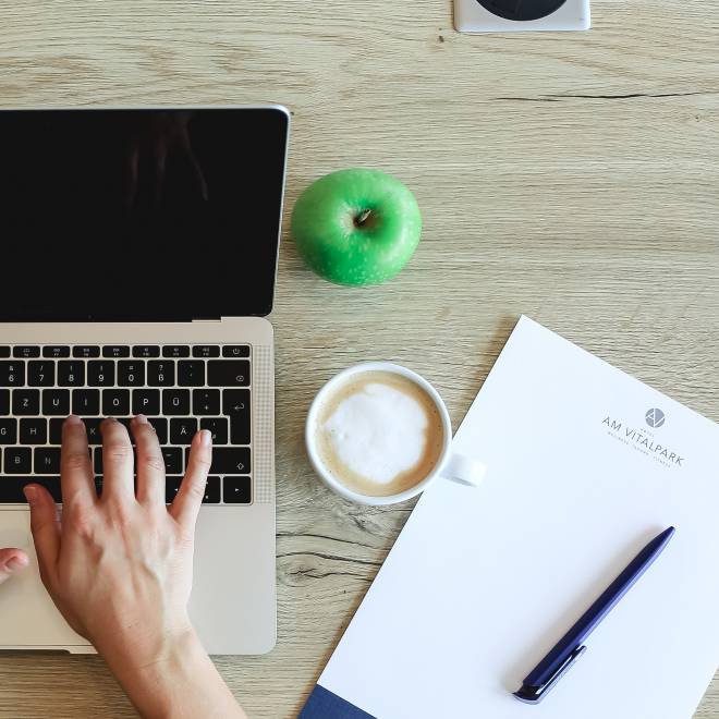 Person schreibt am Laptop mit Kaffee und Block auf dem Tisch